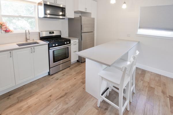 Looking into a bright white kitchen that has two stools pulled up to a white bar height counter in the middle and a wall with a sink and stainless steel appliances on the left. There are also two windows in the room with white shades.