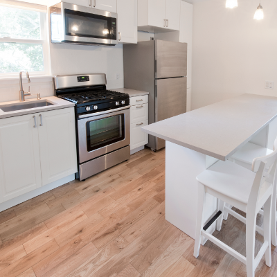 Looking into a bright white kitchen that has two stools pulled up to a white bar height counter in the middle and a wall with a sink and stainless steel appliances on the left. There are also two windows in the room with white shades.