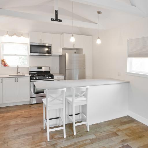 Looking into a bright white kitchen that has two stools pulled up to a white bar height counter in the foreground and a wall with a sink and stainless steel appliances in the back. There are also two windows in the room with white shades.