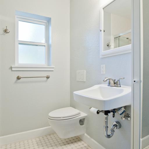 A white tiled bathroom showing a wall with a hanging toilet and sink. There's lighting and a mirror over the sink and a window on the far wall.