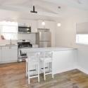Bright and airy kitchen in converted garage Looking into a bright white kitchen that has two stools pulled up to a white bar height counter in the foreground and a wall with a sink and stainless steel appliances in the back. There are also two windows in the room with white shades.