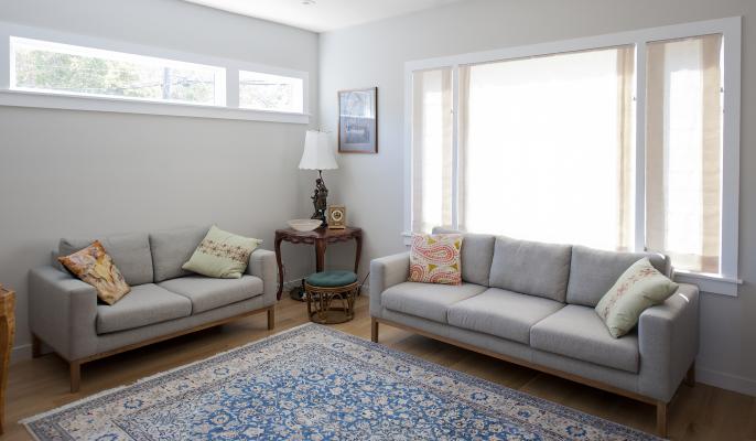 Another angle of the comfortable and modern living room with it's white walls and hardwood floors. There's a gray sofa set and a blue carpet. A row of window on the back wall and a giant window on the right bring in lots of natural light.