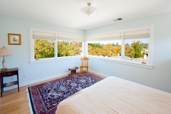 The windows on two walls meet in the corner to provide a stunning, continuous view in the master bedroom. The foot of the bed is in the foreground and a patterned carpet is on the floor. A wooden chair and stool are in the corner. The walls are light blue.
